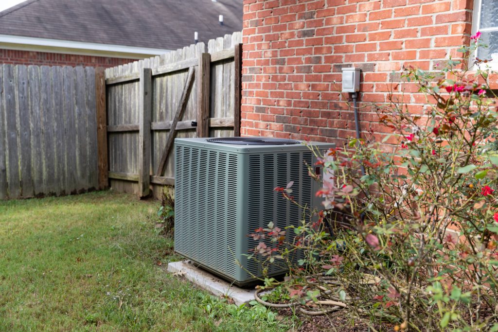 Air conditioner condenser unit sitting next to brick home with fence